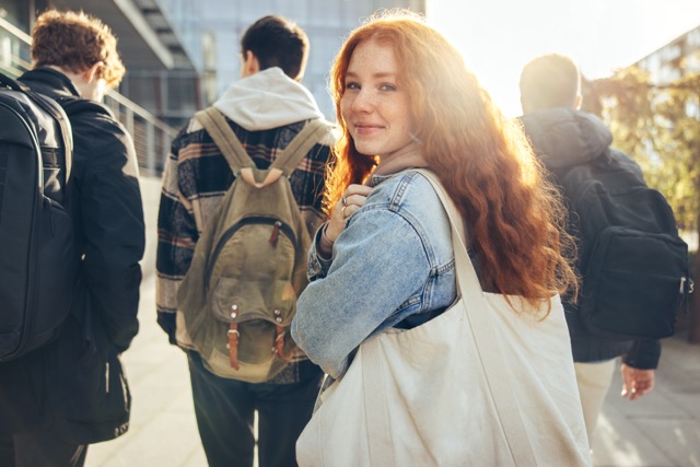 A young woman with long curly red hair and freckles looking back and smiling while walking on a university campus.