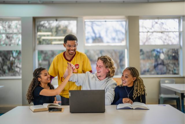 Four university students sitting together at a table indoors, smiling and high-fiving while working on a laptop with open books in front of them, showing collaboration and shared success.