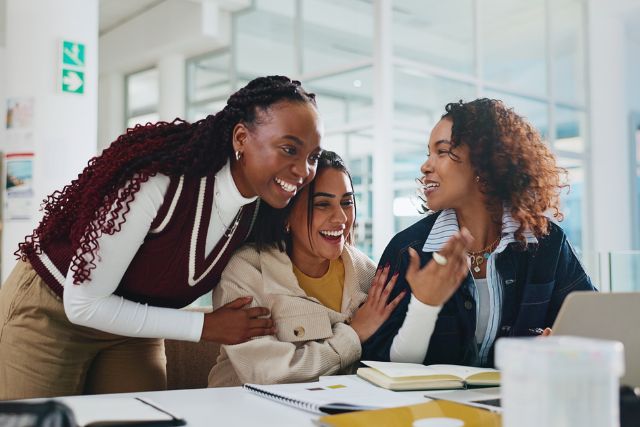 Three female students laughing together while looking at a laptop, illustrating the collaborative community supported by bursaries for 2026.