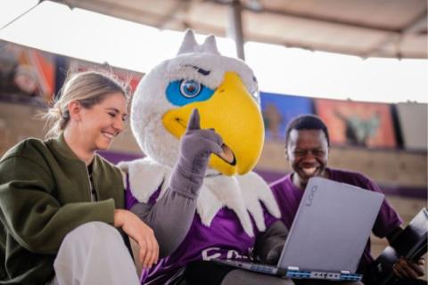 Three NWU students sitting together outdoors with the NWU eagle mascot, smiling and looking at a laptop, representing a fun and supportive student experience on campus.