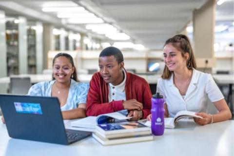 Three NWU students studying together in a library, smiling while looking at a laptop and textbooks, showing collaboration and academic life on campus.