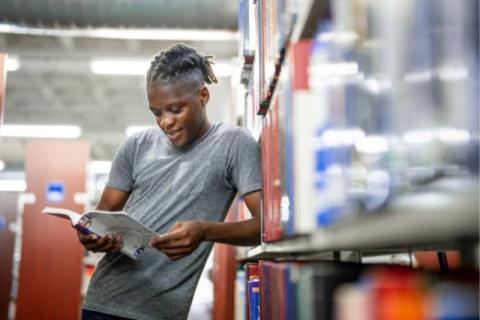 A university student standing between library shelves, leaning against a bookcase while reading a book in a quiet study area.