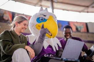 Three NWU students sitting together outdoors with the NWU eagle mascot, smiling and looking at a laptop, representing a fun and supportive student experience on campus.