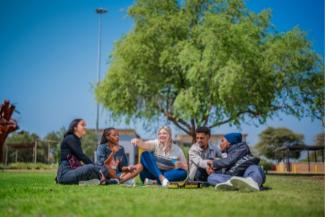Five diverse first-year students sitting on the grass at North-West University campus, laughing and playing a board game under a tree on a sunny day.